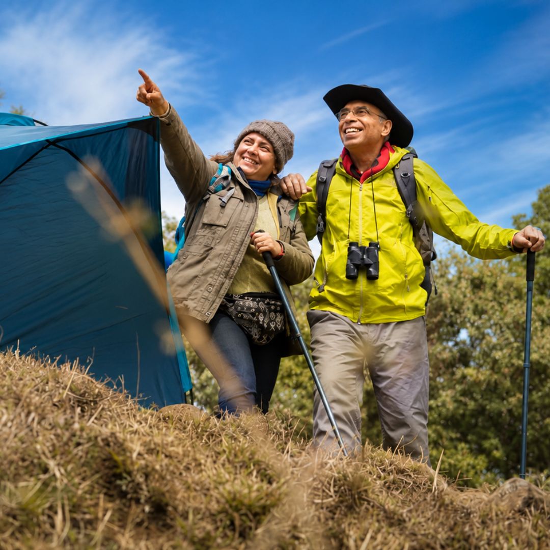 Two hikers with camping gear standing near a tent on a grassy hill with a blue sky.