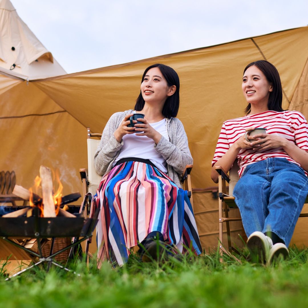 Two women sitting in front of a tent, holding drinks, with a fire pit in the background.