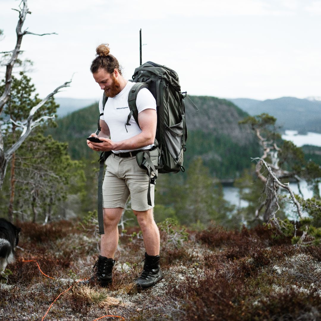 Man with a backpack using a phone in a scenic outdoor setting with mountains and water.