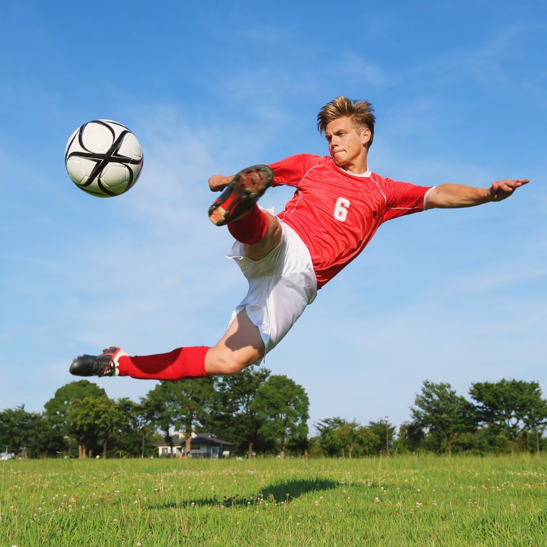 Soccer player in red jersey kicking a ball on a grassy field with blue sky